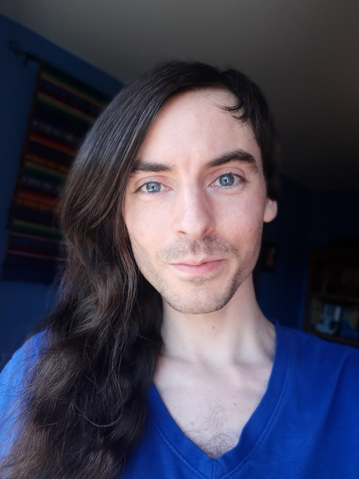 Portrait photo of me. Asymmetrical haircut, long dark brown on one side, short on the other. Large blue eyes. Pale skin. Blue shirt.